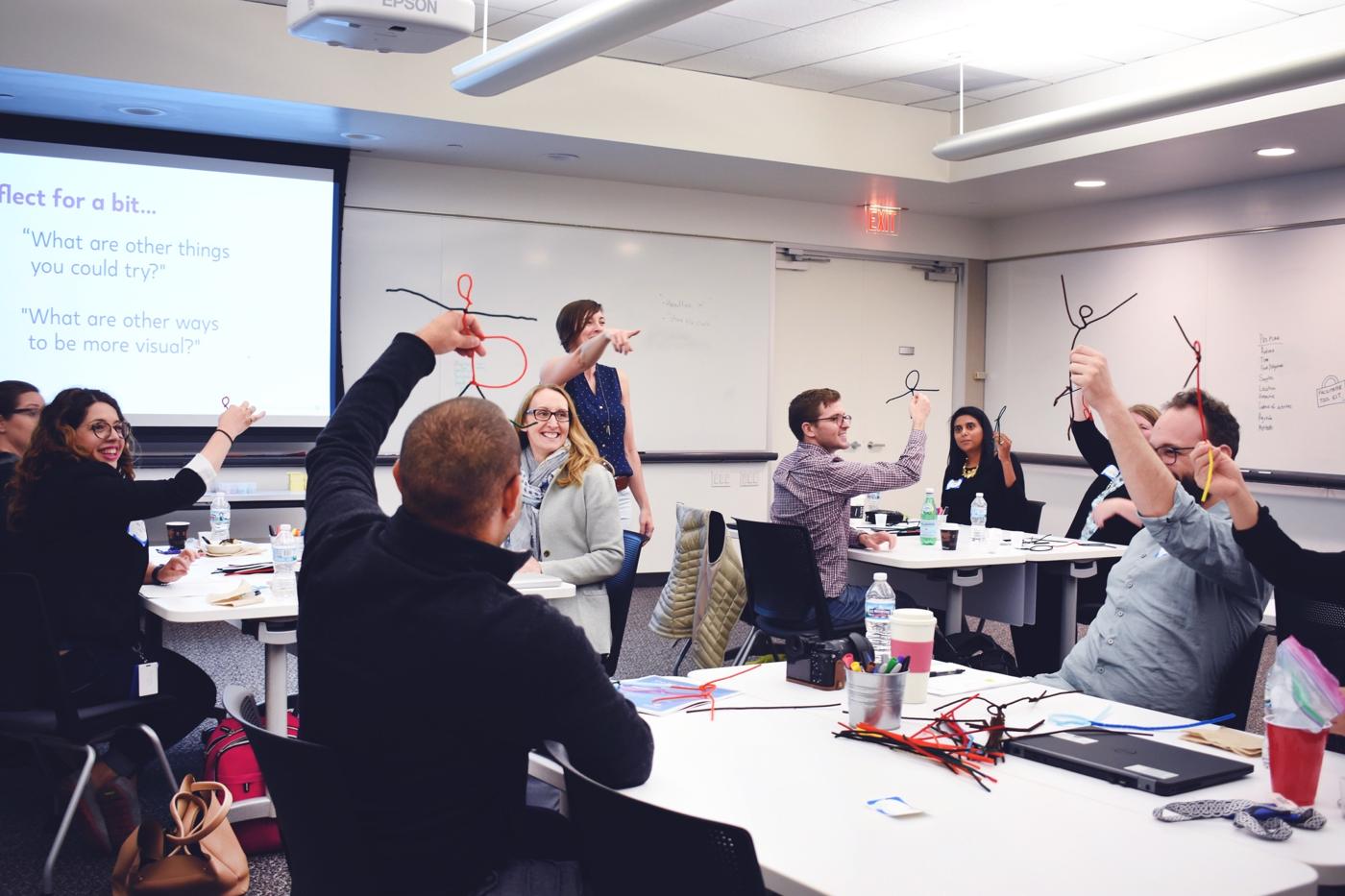Participants in a CXD design thinking workshop holding up pipe-cleaner figures they have made, laughing, around tables covered with materials.