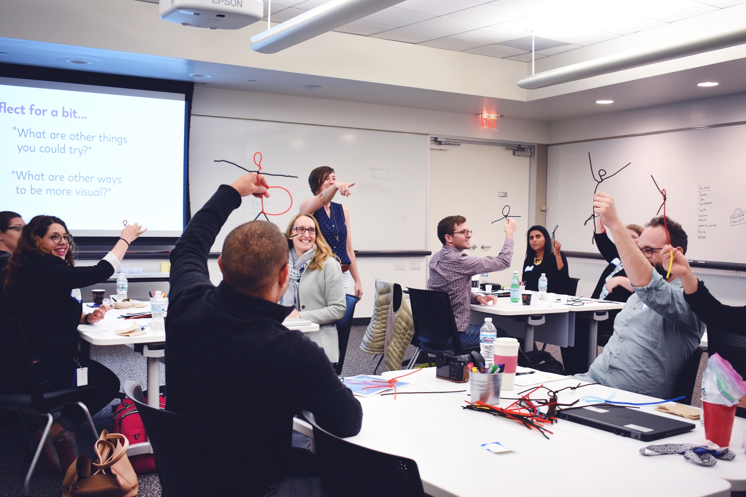 Participants in a CXD design thinking workshop holding up pipe-cleaner figures they have made, laughing, around tables covered with materials.